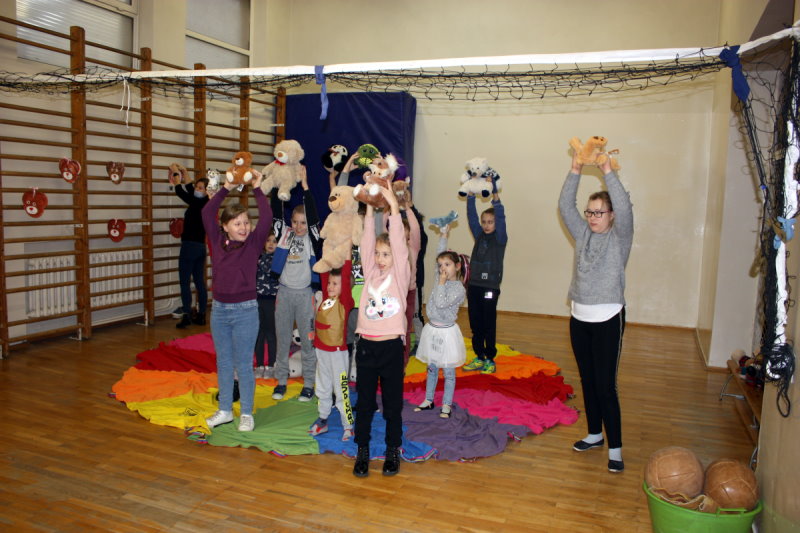Grupa dzieci stoi w sali. Ręce mają podniesione do góry i trzymają w nich maskotki – misie. Na podłodze leży kolorowy materiał. Po prawej stronie piłki w pojemniku.