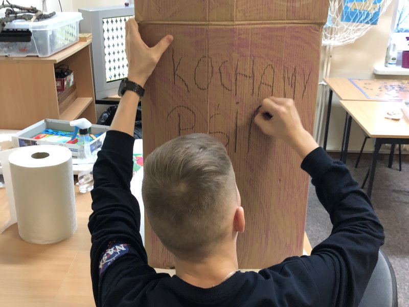 Chłopak pisze na papierowym kartonie w sali. Napis: kochamy ... . W tle ławki, szafka, przybory szkolne i rolka ręcznika papierowego. 