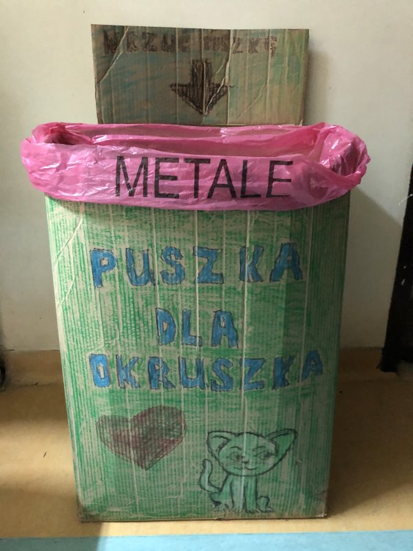 Na stole stoi papierowy karton. Na nim napisy: wrzuć puszkę metale puszka dla okruszka. Jest na nim namalowane serce i kot.