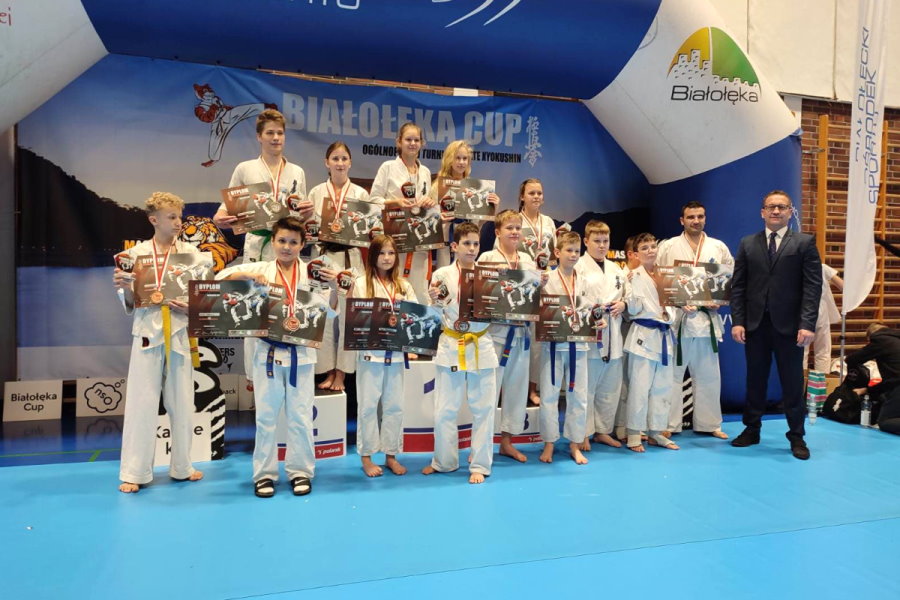 16 osób stoi w sali. Są to zawodnicy karate z trenerem – wspólne zdjęcie. Mają oni na szyi medale a ręku dyplomy. W tle nad nimi napis: Białołęka Cup Ogólnopolski Turniej Karate Kyokushin.