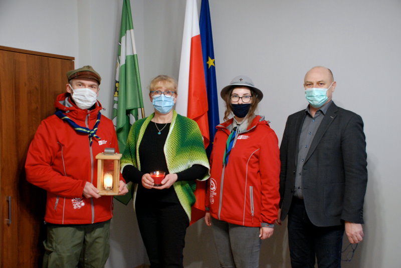 W sali urzędu stoją obok siebie 4 osoby. 2 kobiety i 2 mężczyzn. Jeden z mężczyzn trzyma lampion, a jedna z kobiet świeczkę. Za nimi 3 flagi w stojaku. W tle widać fragment szafki.