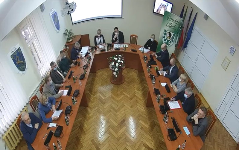 Sala Starostwa Powiatowego w Olecku w czasie sesji. Radni siedzą przy stole w sali urzędu.