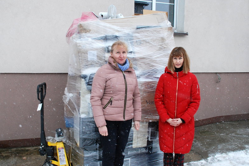 Na podwórku przed budynkiem urzędu stoją obok siebie 2 kobiety. Za nimi jest wysoka sterta kartonów z zużytym sprzętem elektronicznym.