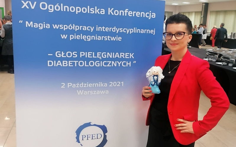 Kobieta stoi przy stojaku promocyjnym w sali. Na nim jest napis: XV Ogólnopolska Konferencja, magia współpracy w pielęgniarstwie, 2 października 2021 r., Warszawa. W ręku trzyma ona figurkę anioła. W tle stoły, a przy nich stoi kilkanaście osób.