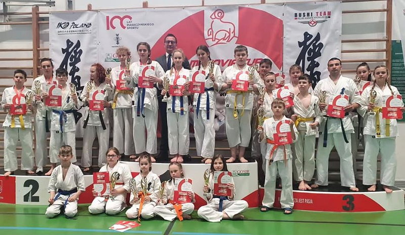 18 osób stoi obok siebie na podium w sali. 5 osób siedzi przed nimi na ziemi. Są to zawodnicy karate z trenerem. Zawodnicy są ubrani w kimona. Trzymają w ręku puchary i dyplomy. Za nimi plakat turnieju.