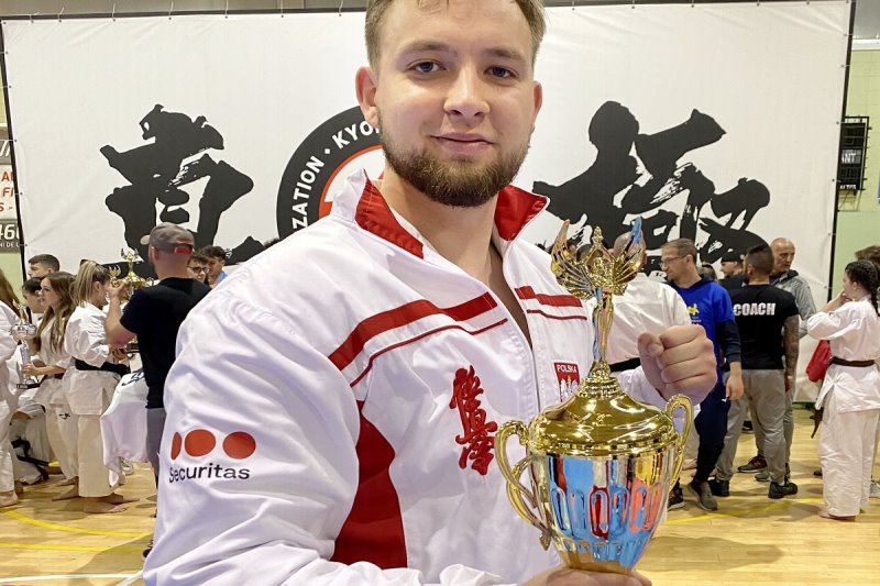 Mężczyzna trzyma w ręku puchar. Za nim stoi kilkanaście osób. Są to zawodnicy karate. Jest też baner zawodów.