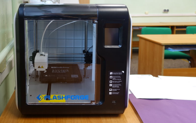 Drukarka 3D stoi na stole w sali. W tle widać ławki z krzesłami.