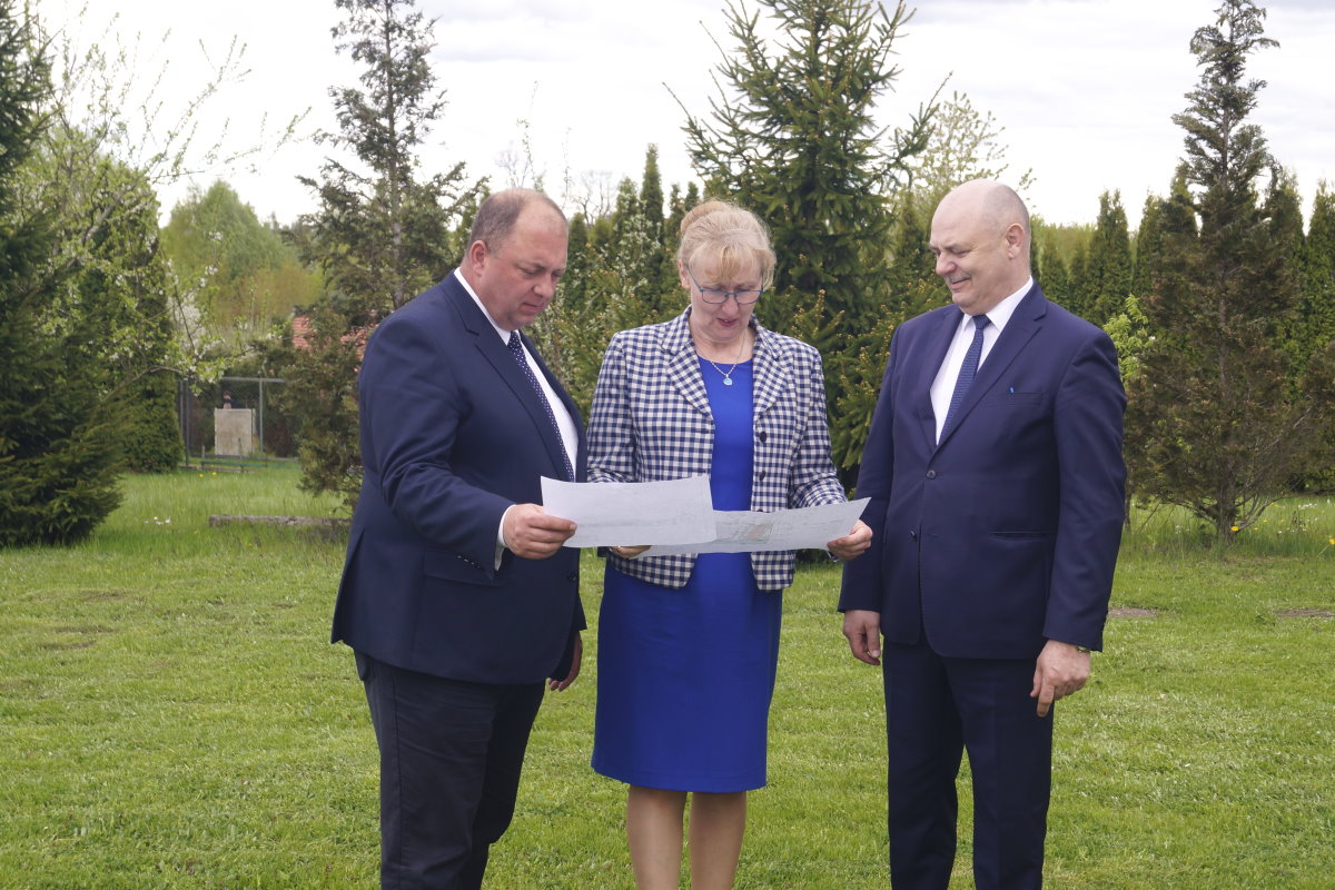 Kobieta i 2 mężczyzn stoi obok siebie na podwórku. Kobieta i jeden z mężczyzn trzymają w rękach kartki. Wszyscy patrzą na nie. W tle drzewa i rośliny.