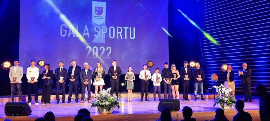 17 osób stoi na scenie w sali. Trzymają oni statuetki. Za nimi napis: Gala Sportu 2022 i logo urzędu. Na scenie są też kwiaty w 2 doniczkach.