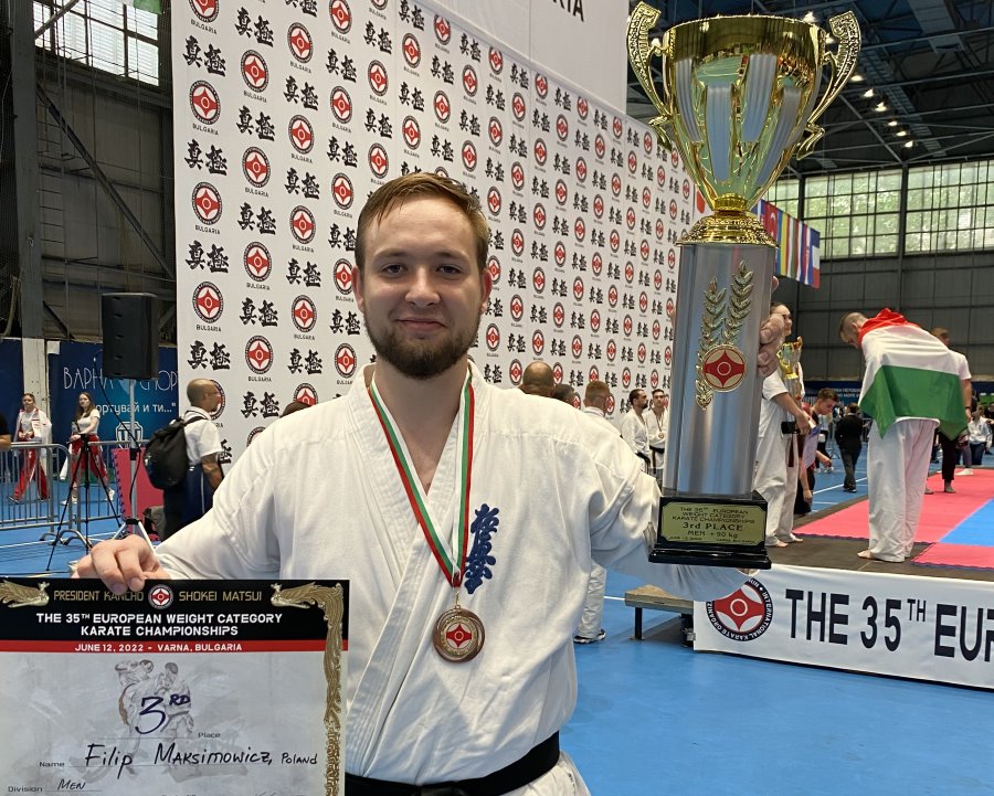 Mężczyzna stoi w sali. Jest to zawodnik karate. Trzyma on puchar i dyplom. Na szyi ma medal. Za nim stoi kilkanaście osób, tj. zawodników karate i banery zawodów.