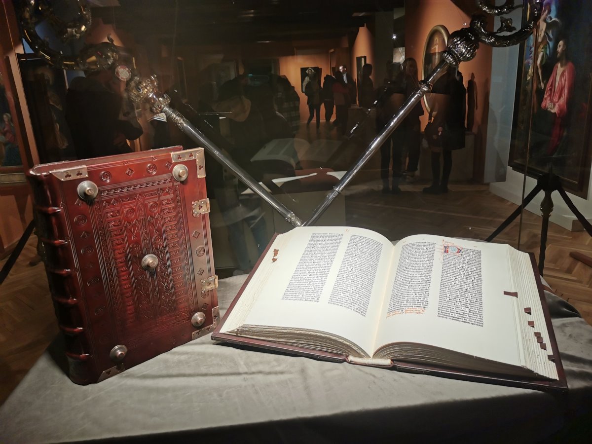 Sala w muzeum. W gablocie leży otwarta księga. Obok niej stoi księga w ozdobnej okładce. W tle eksponaty i kilka ludzi, którzy stoją w tej sali.