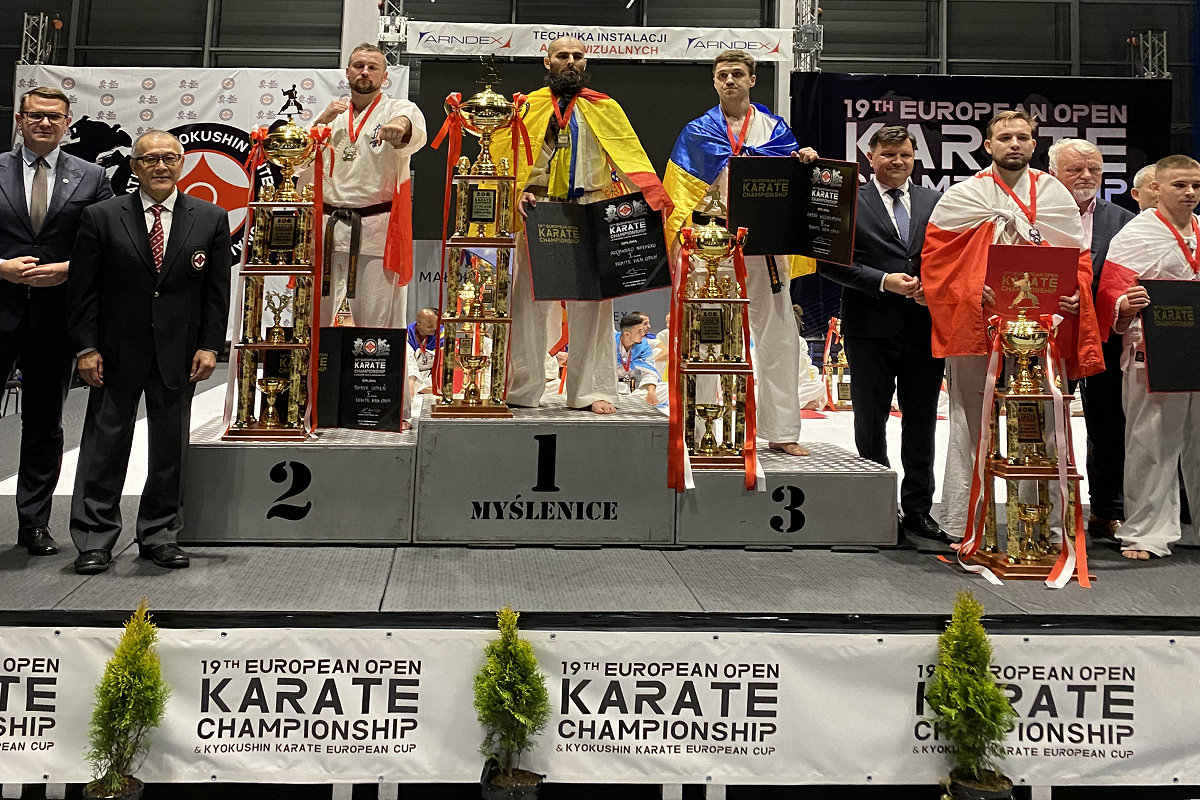 3 zawodników na podium. Obok nich 4 mężczyzn i 2 zawodników. Są to zawodnicy karate i są ubrani w kimona. Trzymają oni puchary i dyplomy. Mają zawiązane na plecach i szyi flagę swojego kraju. Za nimi banery promocyjne.