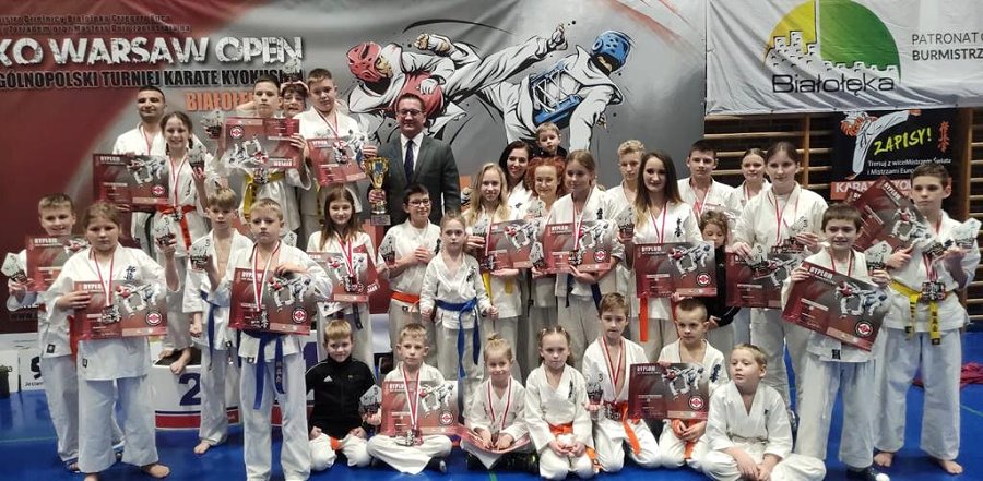 W sali stoi obok siebie 26 osób. Przed nimi siedzi na podłodze 7 osób. Wszyscy, oprócz 1 mężczyzny, są ubrani w kimono. Są zawodnicy karate z trenerem. Większość z nich trzyma puchary i dyplomy. Ma medale na szyi. Za nimi baner z nazwą zawodów karate.