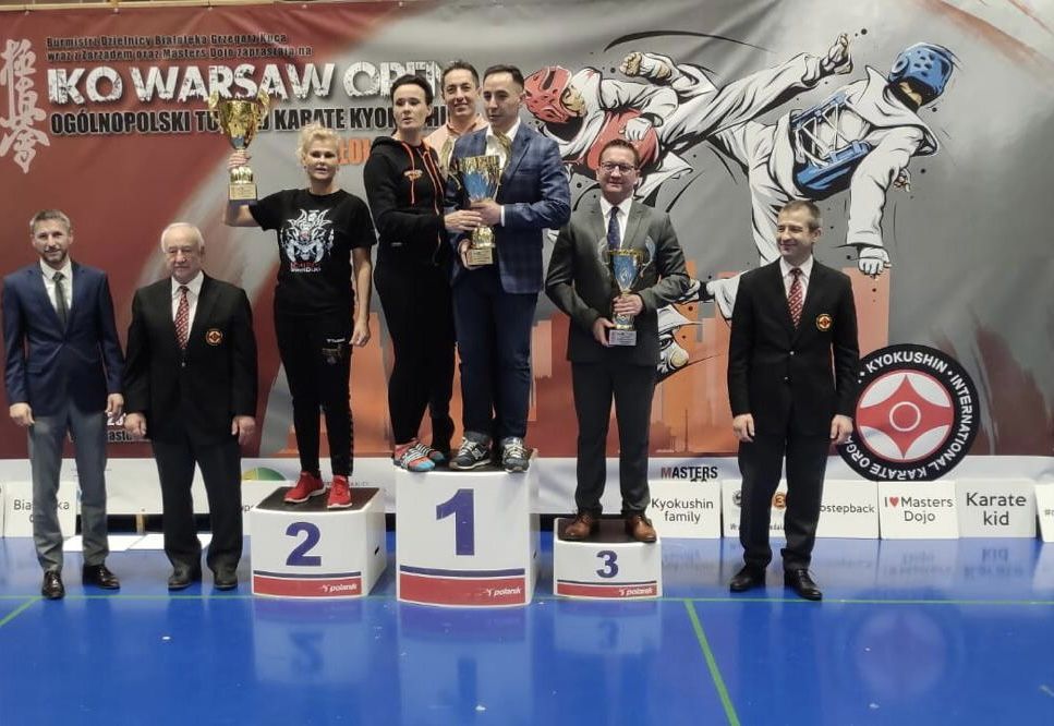 8 osób stoi obok siebie w sali, tj. 2 kobiety i 6 mężczyzn. 5 z tych osób stoi na podium. Trzymają oni puchary. Za nimi baner z nazwą zawodów karate.