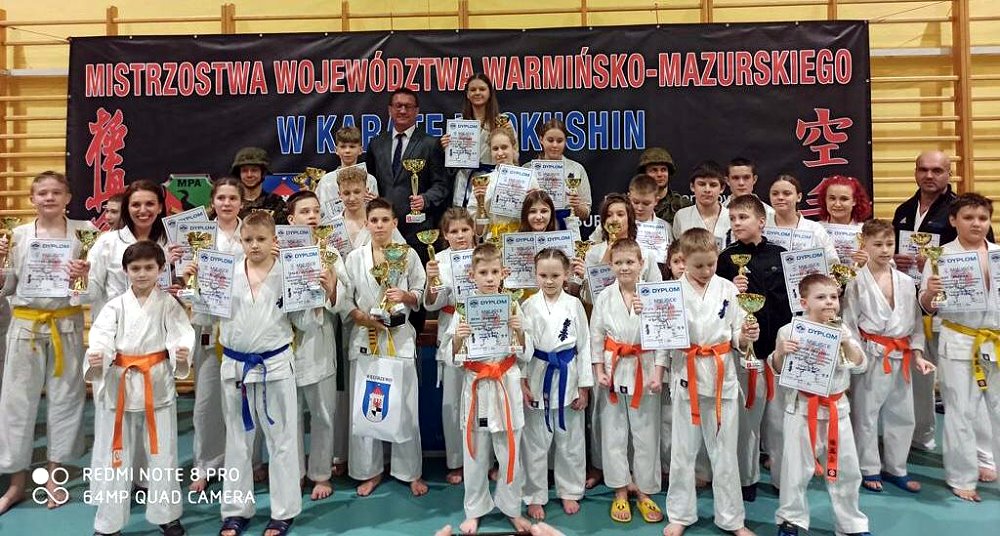 Kilkadziesiąt osób stoi obok siebie w sali w budynku. Są to dziewczęta, chłopcy, kobieta i mężczyźni. Większość z nich to zawodnicy karate. Są oni ubrani w stroje sportowe, tj. kimona. Trzymają oni puchary i dyplomy. Za nimi baner z nazwą zawodów.