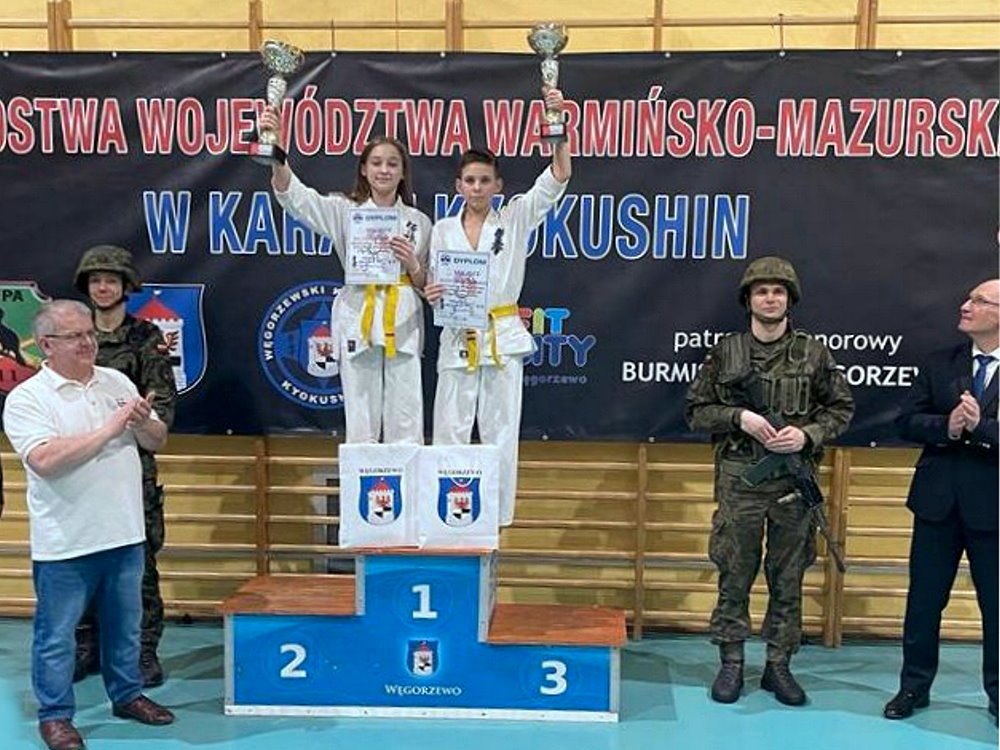 Dziewczyna i chłopak stoją obok siebie na podium w sali w budynku. Są to zawodnicy karate. Są oni ubrani w stroje sportowe, tj. kimona. Trzymają oni puchary i dyplomy. Po obu ich stronach stoi po 2 osoby. Za nimi baner z nazwą zawodów.
