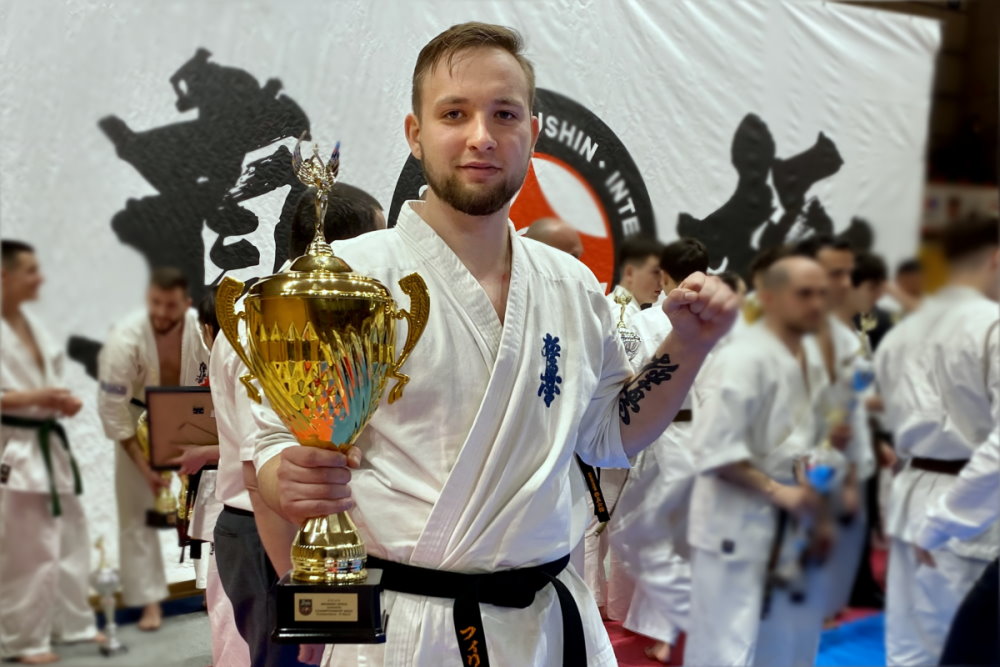 Mężczyzna stoi w sali i trzyma w dłoni puchar. Jest to zawodnik karate. Jest on ubrany w kimono. Za nim baner zawodów i kilkanaście osób – zawodników karate.