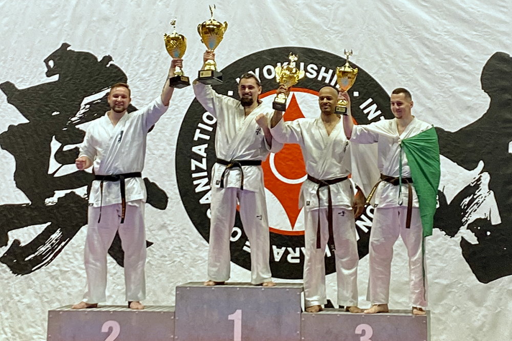 4 mężczyzn stoi obok siebie w sali na podium. Trzymają oni w dłoni puchary. Są to zawodnicy karate. Są oni ubrani w kimono. Za nim baner zawodów.