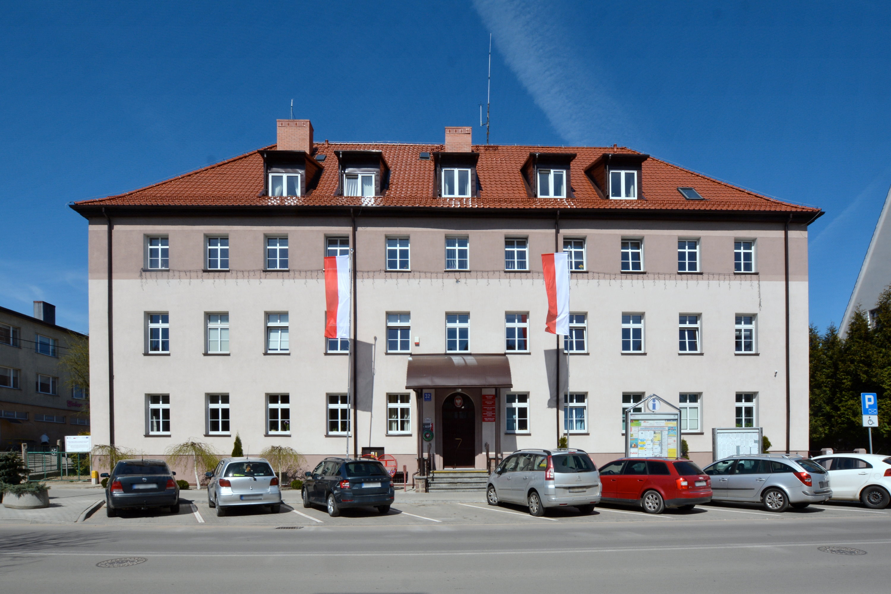 Budynek. Przed nim 2 flagi Polski na masztach, 2 tablice informacyjne, chodnik, parking, znak drogowy i ulica. Na parkingu jest 7 samochodów. Po lewej budynek i drzewa, a po prawej drzewa.