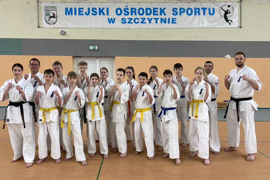 17 osób stoi obok siebie w budynku. To jest: 3 mężczyzn, kobieta, 8 chłopaków i 5 dziewczyn. Są to zawodnicy karate. Są ubrani w kimona. Za nimi ściana. Na niej baner z napisem: Miejski Ośrodek Sportu w Szczytnie i 2 logotypy po bokach.