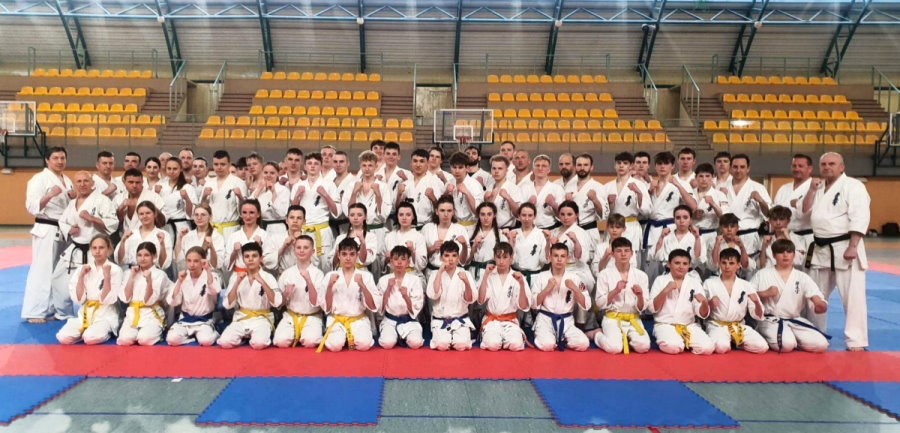 Kilkadziesiąt osób obok siebie w budynku. Są to mężczyźni, kobiety, chłopacy i dziewczyny. Są to zawodnicy karate. Są ubrani w kimona. Niektórzy z nich stoją, a niektórzy siedzą na podłodze. Za nimi trybuny z siedzeniami.
