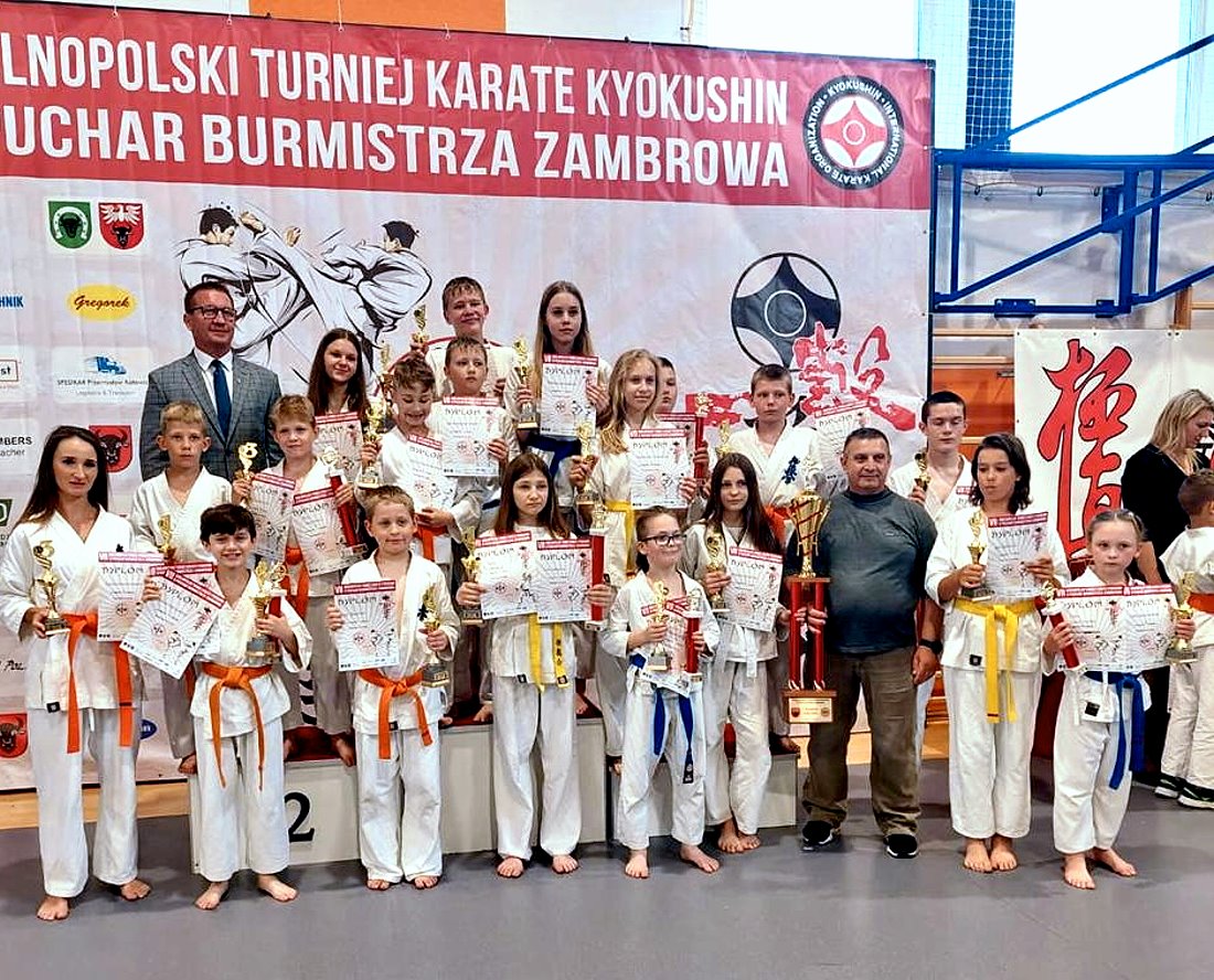 W sali w budynku kilkadziesiąt osób stoi obok siebie. Jest to młodzież i 2 mężczyzn, kobieta. Młodzież trzyma dyplomy i puchary. Są oni ubrani w kimona. Za nimi baner z napisem: Ogólnopolski Turniej Karate Kyokushin o Puchar Burmistrza Zambrowa.