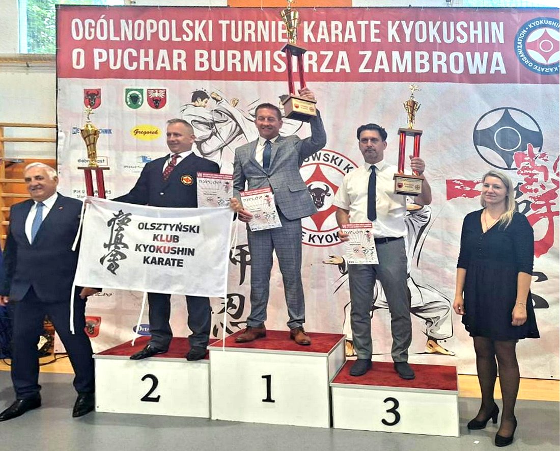 Na podium 3 mężczyzn z dyplomami i pucharami. Ten z podium nr 2 ma na materiale napis: Olsztyński Klub Kyokushin Karate. Po bokach mężczyzna i kobieta. Za nimi baner z logotypami i napis: Ogólnopolski Turniej Karate Kyokushin o Puchar Burmistrza Zambrowa.
