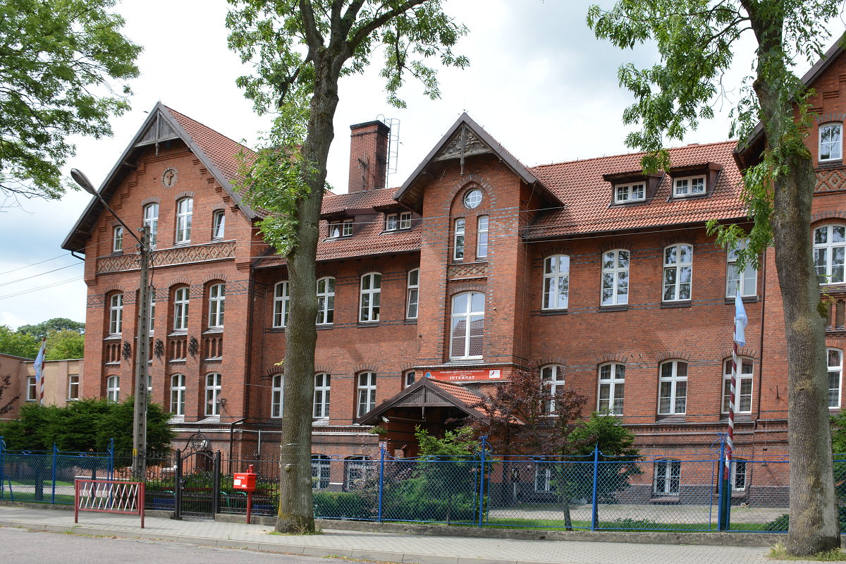 Budynki. Jest to internat szkoły - Ośrodka Szkolno-Wychowawczego dla Dzieci Głuchych w Olecku. Przed nimi jest chodnik, trawa, krzewy i drzewka. Za nimi ogrodzenie z wejściem, drzewa, skrzynka na listy, chodnik i ulica.