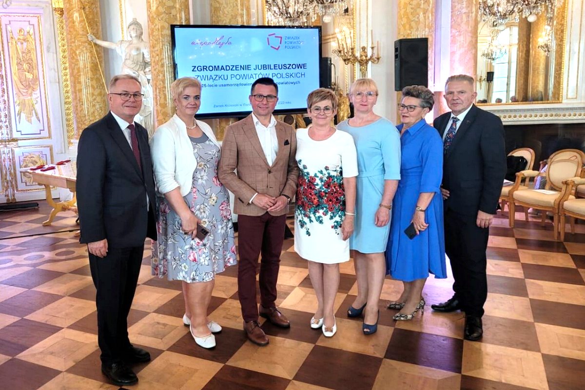 W sali stoją obok siebie 4 kobiety i 3 mężczyzn. 2 z nich trzymają telefony komórkowe. Za nimi ekran, na którym są wyświetlone 2 logotypy i napis: Zgromadzenie Jubileuszowe Związku Powiatów Polskich. Za nimi fotele, rzeźby, lustra, świeczniki.