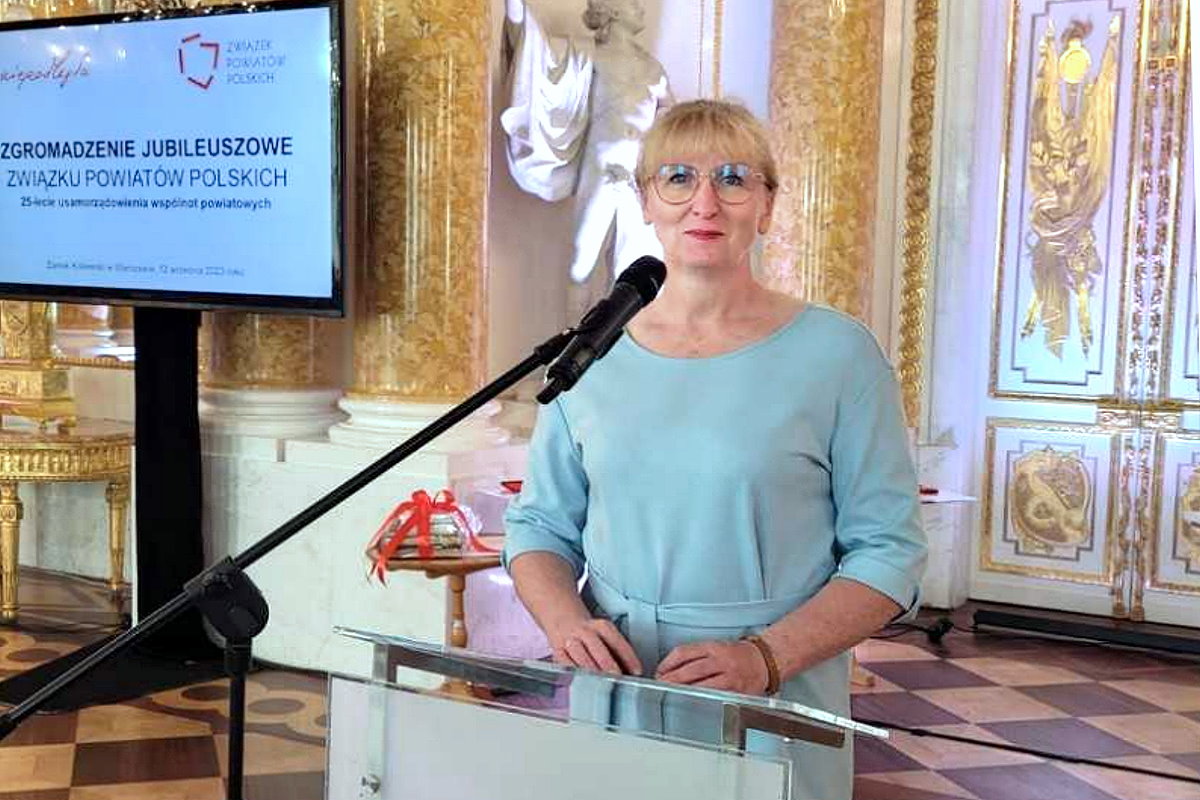 W sali przy mównicy z mikrofonem stoi kobieta. Za nią ekran. Na nim są wyświetlone 2 logotypy i napis: Zgromadzenie Jubileuszowe Związku Powiatów Polskich. W tle ściany ze zdobieniami i rzeźby.