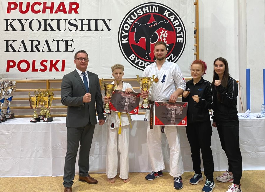 5 osób stoi w sali. 3 z nich to zawodnicy karate, a 2 z nich to trenerzy – wspólne zdjęcie. 2 zawodników trzyma w rękach puchary i dyplomy. W tle napis: Puchar Karate Kyokushin Polski i stół, na którym stoją puchary.