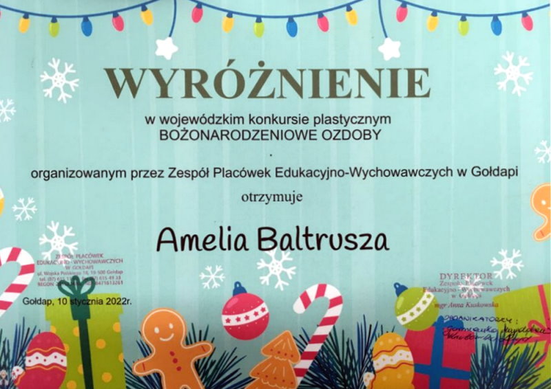 Dyplom z ozdobami świątecznymi. Na nim napis: wyróżnienie w wojewódzkim konkursie plastycznym Bożonarodzeniowe ozdoby, organizowanym przez Zespół Placówek Edukacyjno-Wychowawczych w Gołdapi, otrzymuje Amelia Baltrusza. Gołdap, 10.01.2022 r. 2 pieczątki.