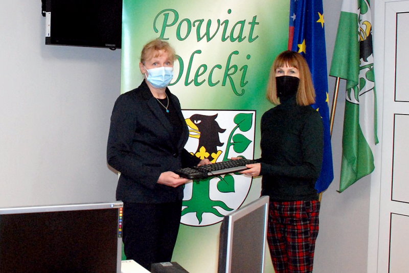2 kobiety stoją w sali. Przekazują sobie klawiaturę. Za nimi stojak z herbem i napisem Powiat Olecki oraz flagi w stojaku. W tle telewizor na ścianie i monitory komputerów.