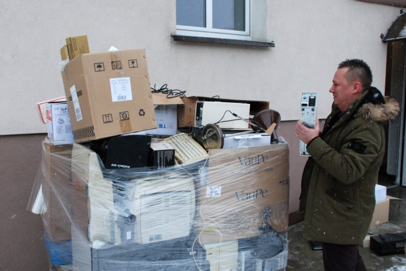 Na podwórku przed budynkiem urzędu stoi mężczyzna. Kładzie rzecz na wysoką stertę kartonów z zużytym sprzętem elektronicznym.