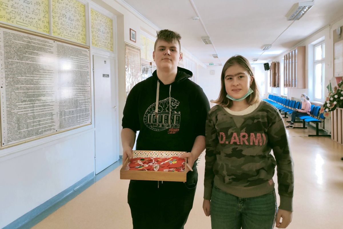 Chłopak z dziewczyną stoją obok siebie na korytarzu w szkole. On trzyma pudełko z sercami. W tle 3 osoby. 2 z nich idą po schodach a 1 – siedzi na krześle. Na ścianach są tablice i gabloty.