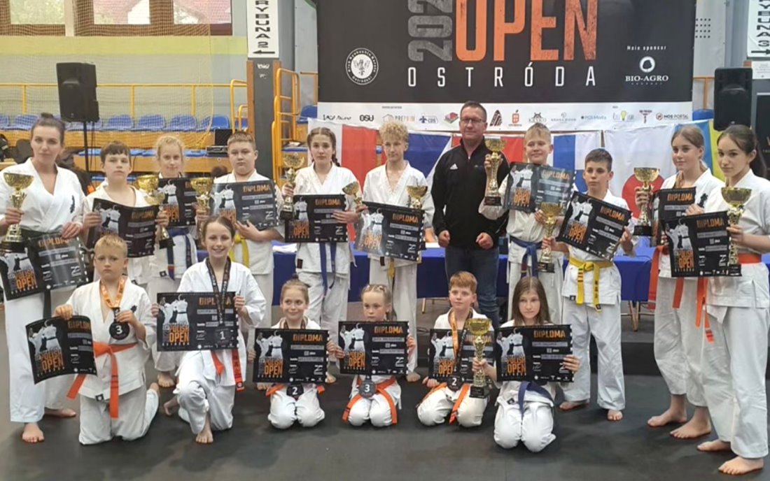 17 osób w sali. W tym 11 osób stoi obok siebie, a przed nimi przykucnęło 6 osób. Są to zawodnicy karate z trenerem. Zawodnicy są ubrani w kimona i trzymają w rękach puchary i dyplomy. Za nimi banery.