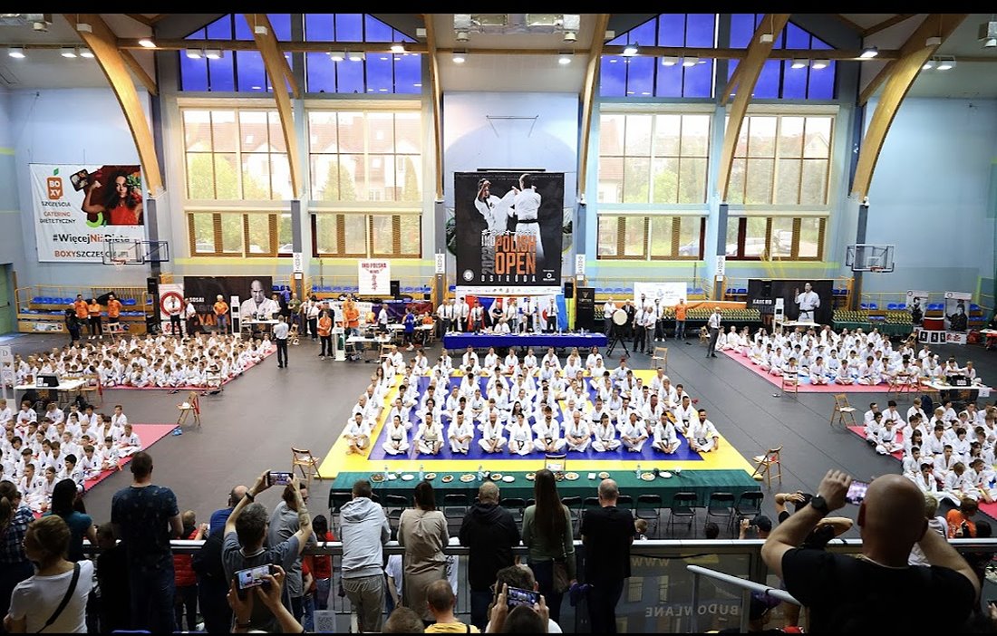 Sala sportowa. Siedzą w niej na podłodze zawodnicy karate. Siedzą oni w 5 grupach. Są oni ubrani w kimona. W tle okna, banery i ludzie, którzy na nich patrzą.