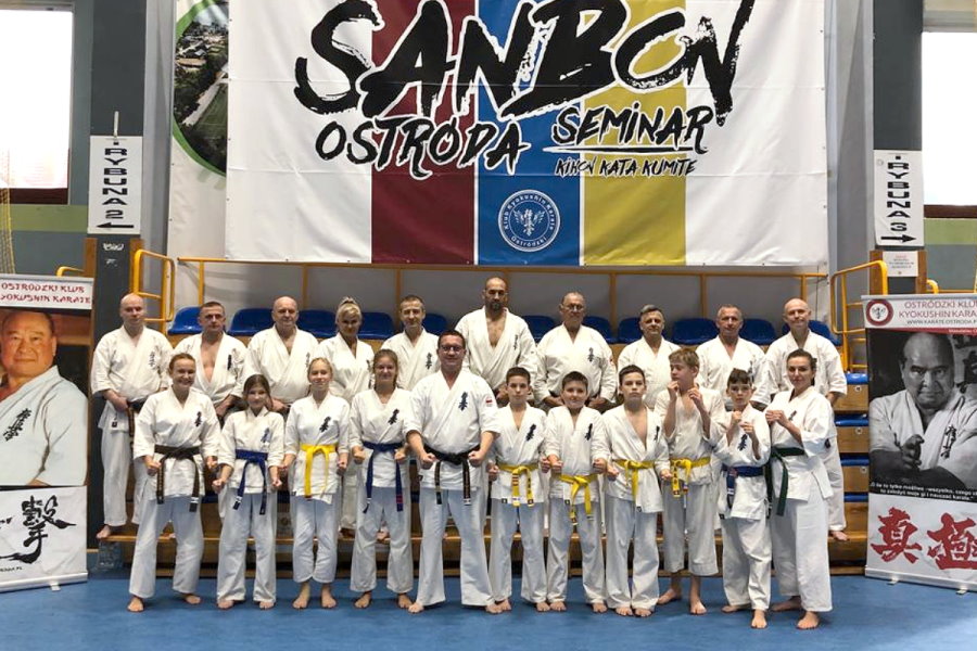21 osób stoi w sali w budynku, tj. 10 mężczyzn, 3 kobiety, 5 chłopaków i 3 dziewczyny. Są to zawodnicy karate. Są oni ubrani w kimona. Za nimi baner z logo i nazwą seminarium. Po bokach stojaki z nazwą klubu i zdjęciami karateków. 