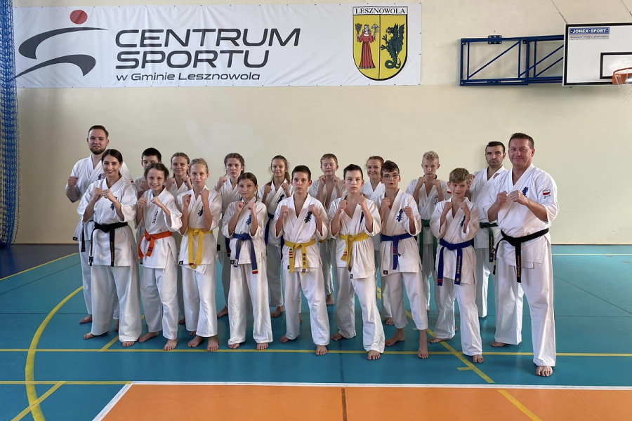18 osób stoi w sali w budynku, tj. 3 mężczyzn, kobieta, 7 chłopaków i 7 dziewczyn. Są to zawodnicy karate. Są oni ubrani w kimona. Za nimi baner z logo i napisem: Centrum Sportu w gminie Lesznowola i herbem urzędu.