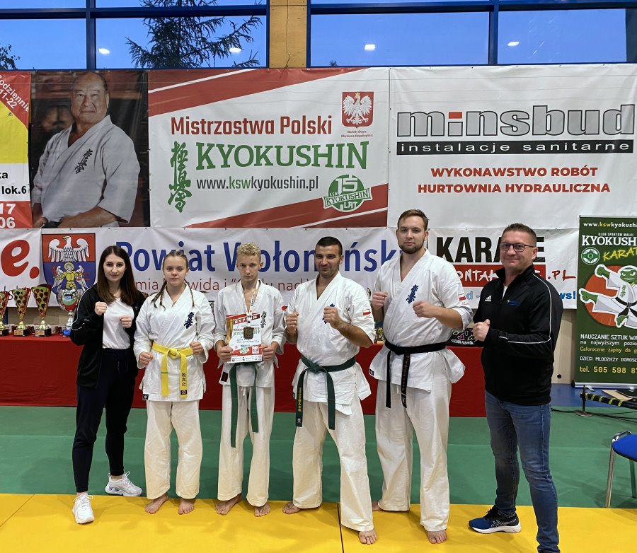 6 osób stoi obok siebie w sali, tj. kobieta, dziewczyna i 3 mężczyzn. Są to zawodnicy karate z trenerami. Za nimi banery promocyjne i puchary.