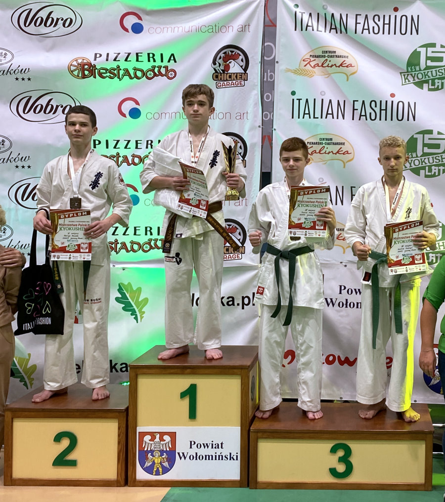 4 chłopaków stoi obok siebie na podium w sali. Są to zawodnicy karate z trenerami. Za nimi banery promocyjne.