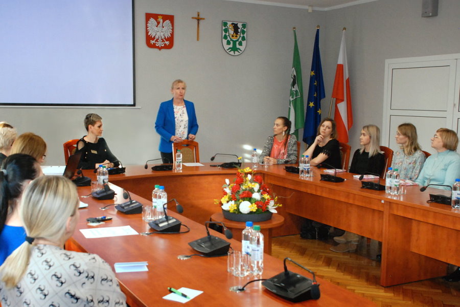 10 kobiet jest w sali w urzędzie. 9 z nich siedzi przy stole, a 1 stoi. Na stole są mikrofony, kartki, długopisy, butelki z wodą i szklanki. Przed stole jest wiązanka z kwiatów. Za nimi stojak z 3 flagami, a na ścianie godło Polski, krzyż i herb urzędu.