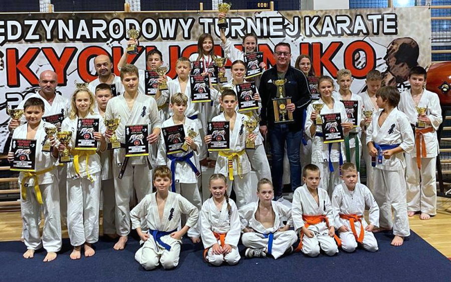 20 osób stoi obok siebie w sali. Przed nimi siedzi na podłodze 5 osób. Są to dziewczyny, chłopaki i mężczyźni. To zawodnicy karate z trenerem. Zawodnicy są ubrani w kimono. Większość z nich trzyma puchary i dyplomy. Za nimi baner z nazwą zawodów.