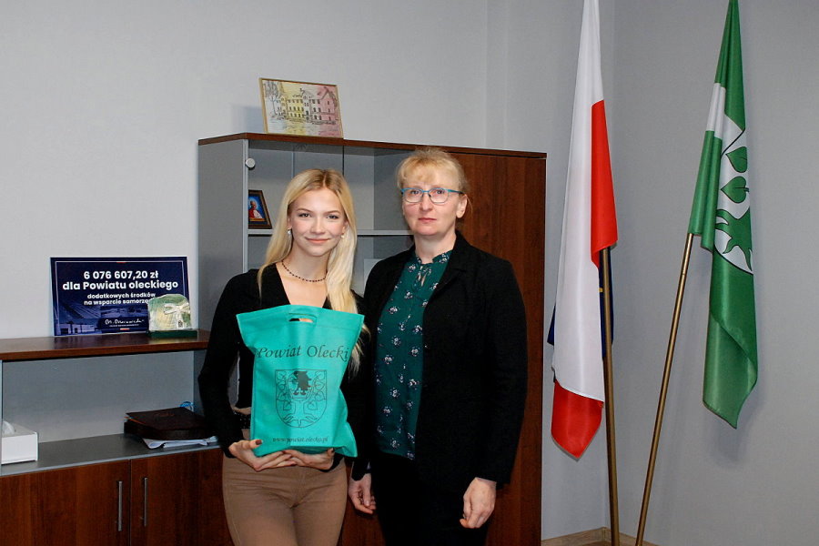 2 kobiety stoją obok siebie w pokoju w urzędzie. Jedna z nich trzyma torebkę z herbem i napisem: Powiat Olecki. Za nimi stoją szafki z półkami i różnymi przedmiotami oraz stojak z flagami.