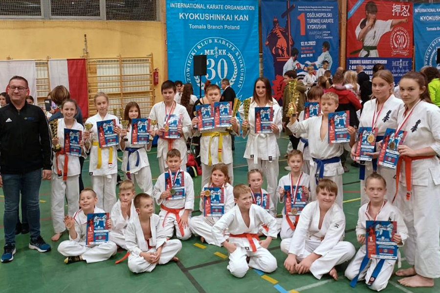 Mężczyzna, 6 dziewczyn i 4 chłopców stoi obok siebie w sali. Przy nich na podłodze siedzi 8 chłopaków i 2 dziewczyny. To zawodnicy karate z trenerem. Większość zawodników ma w rękach dyplomy i puchary. Są oni ubrani w kimono. Niektórzy mają medale na szyi.