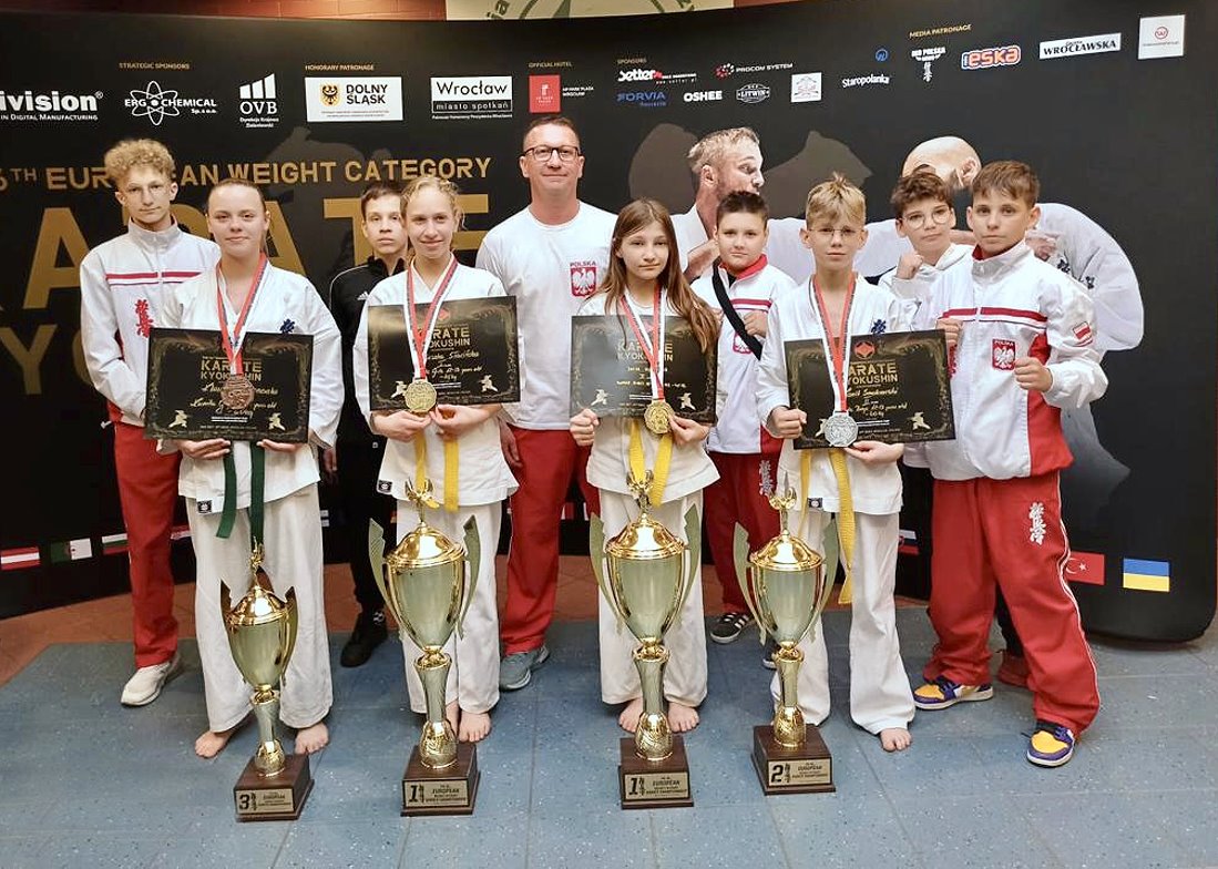 3 dziewczyny, 6 chłopaków i mężczyzna stoją obok siebie w budynku. Są oni ubrani w sportowe stroje. Dziewczyny i jeden z chłopaków mają dyplomy i medale na szyi. Przed nimi stoją 4 puchary. Za nimi wszystkimi jest stojak z logami i nazwą zawodów.
