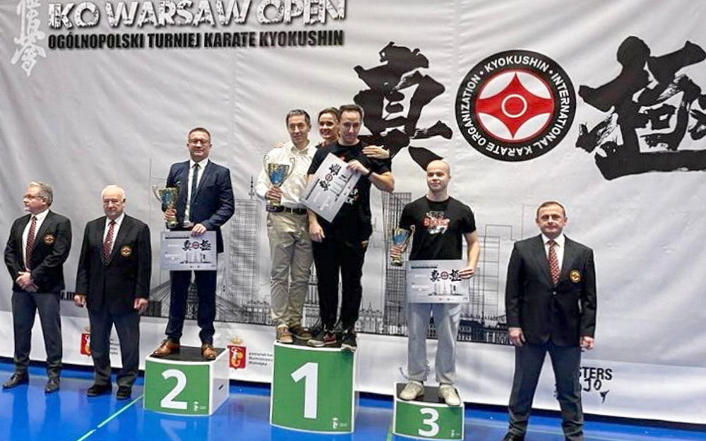 W sali na podium z nr 2 stoi mężczyzna, a na podium z nr 3 – chłopak, z nr 1 – 2 mężczyzn i kobieta. Trzymają oni dyplomy i puchary. Po jednej ich stronie stoi 2 mężczyzn, a po drugiej 1 mężczyzna. W tle baner promocyjny z nazwą zawodów.