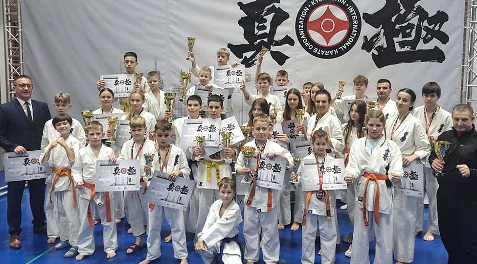 W sali obok siebie stoi około 30 osób. Są to chłopaki, dziewczyny, mężczyźni i kobiety. Jeden z chłopaków przy nich przykucnął. Jeden z mężczyzn jest ubrany w garnitur, a drugi w dresy. Pozostali są w kimonach. Większość z nich trzyma dyplomy i puchary.