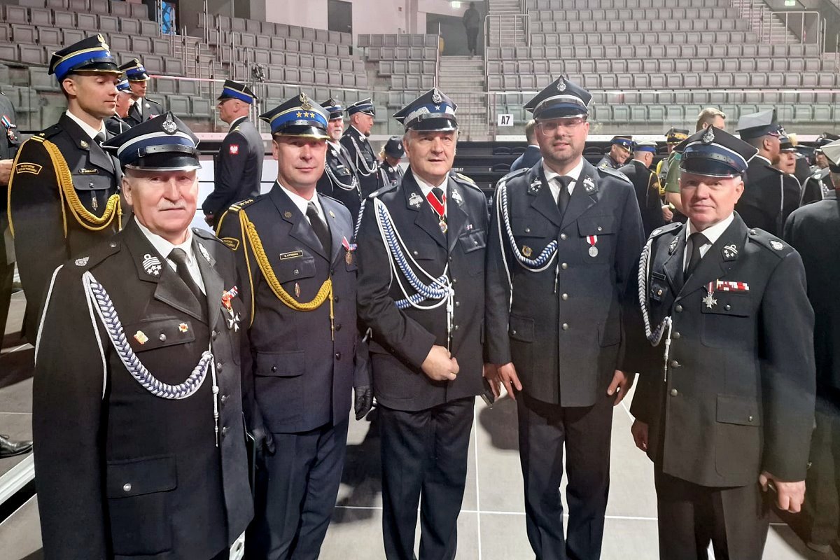 W budynku stoi obok siebie 5 mężczyzn. Są oni ubrani w wyjściowe mundury strażackie. Za nimi kilkanaście strażaków w mundurach i kilkadziesiąt krzesełek na trybunach.