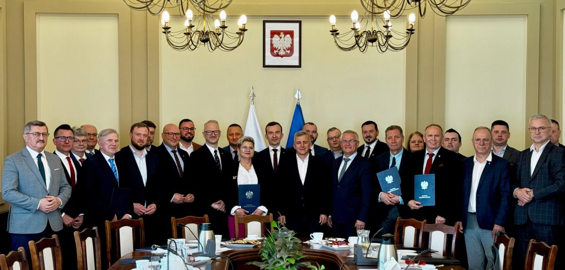 W pomieszczeniu 24 mężczyzn i 2 kobiety stoją obok siebie. Jedna z kobiet i 2 mężczyzn trzymają okładki z herbem. Za nimi 2 flagi i godło Polski na ścianie. Przed nimi stół z różnymi przedmiotami.